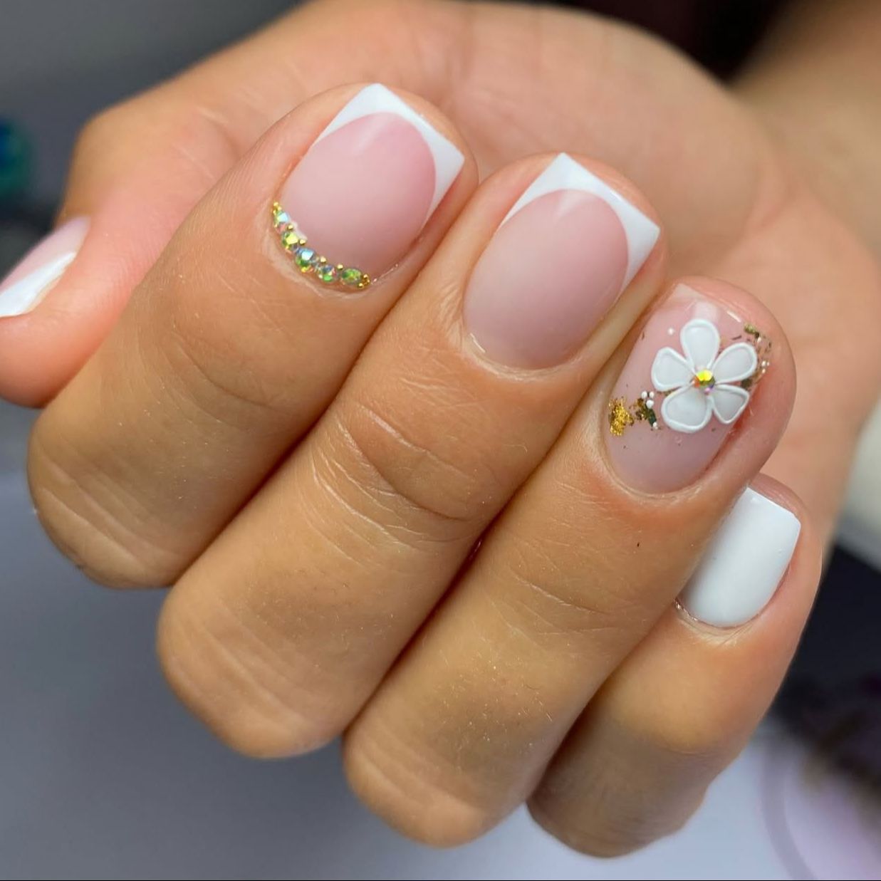 Manicura francesa blanca sobre base nude con una flor blanca y pedrería dorada